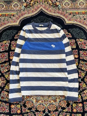 Randig långärmad topp Abercrombie - Snygg långärmad topp från Abercrombie & Fitch med breda blå och vita ränder. Klassisk rund halsringning och broderad logga på bröstet. Perfekt för en avslappnad och trendig stil.