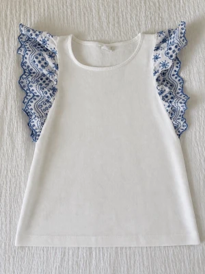 Vit topp med broderade volangärmar - Superfin vit topp med breda volangärmar i vitt och blått broderi. Ärmlös modell med rund halsringning och detaljerade broderier på ärmarna som ger en romantisk vibe. Perfekt till jeans eller kjol för en söt look.