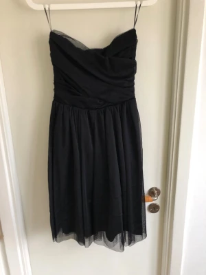 Svart axelbandslös festklänning CUBUS - Snygg svart axelbandslös klänning med omlottliv och lätt utställd kjol i tunt, transparent tyg. Perfekt för dig som gillar en klassisk och stilren look. Klänningen har en feminin siluett och tunna axelband som kan tas av.