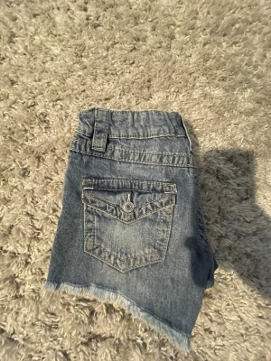Blå jeansshorts från Gina Tricot - Snygga blå jeansshorts från Gina Tricot med rå kant nertill och fin bakficka med knapp. Shortsen har bälteshällor och dragkedja framtill. Perfekt nu till sommaren som matchar med allt