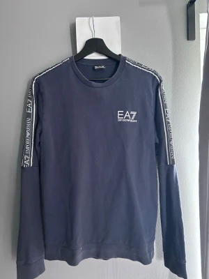 Mörkblå EA7 sweatshirt med logga - Snygg mörkblå sweatshirt från EA7 Emporio Armani med vita logotexter längs ärmarna och tryckt logga på bröstet. Tröjan har rund halsringning och långa ärmar. Perfekt för en avslappnad och trendig stil.