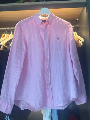 Ljusrosa linneskjorta från Polo Ralph Lauren - Säljer en ljusrosa skjorta från Polo Ralph Lauren i linne med klassisk button-down krage och broderad logga på bröstet. Skjortan har långärmad design och knappar framtill. Perfekt för dig som gillar stilrena plagg med en touch av färg. Snygg och stek som fon inför sommaren🌻🕶️⛱️🍉