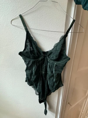 Mörkgrön spetsbody med smala axelband - Snygg mörkgrön body från NA-KD med spetsdetaljer och smala justerbara axelband. Bodyn har v-ringning, genomskinliga partier och dekorativa sömmar. Materialet är en kombination av mesh och spets som ger en lyxig känsla.