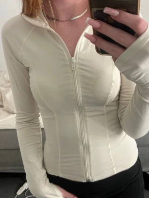 Vit långärmad ziptop - Snygg vit långärmad topp med dragkedja framtill och figurnära passform. Toppen har tumhål i ärmsluten och en hög krage. Perfekt för en trendig och sportig look.