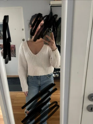 Vit stickad tröja med v-ringning - Säljer en krämvit stickad tröja med djup v-ringning och långa ärmar. Tröjan har en chunky ribbstickad struktur och avslappnad passform, perfekt att styla med jeans. Materialet ser ut att vara mjukt och mysigt, troligen bomull eller akryl.