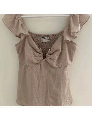 Beige volangtopp från Aware - Supersöt beige topp från Aware med volangdetaljer vid axlarna och v-ringning med små pärlor framtill. Toppen är ärmlös och har en lätt och luftig känsla, perfekt för varma dagar.