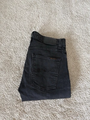 Svarta jeans från Nudie Jeans - Säljer ett par svarta jeans från Nudie Jeans med klassisk femficksdesign och raka ben. Jeansen har en diskret logga på bakfickan och är tillverkade i mjuk denim med lite stretch för extra komfort.