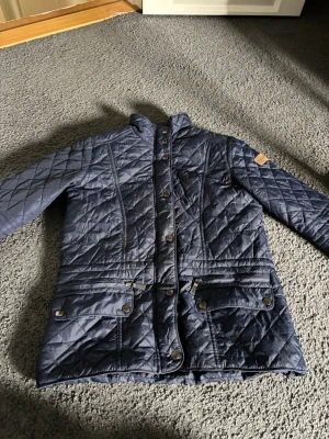 Mörkblå quiltad jacka med knappar - Snygg mörkblå quiltad jacka med lång ärm och hög krage. Jackan har tryckknappar framtill, dragkedja och flera fickor med lock. Perfekt för kyligare dagar och har en klassisk, tidlös look.