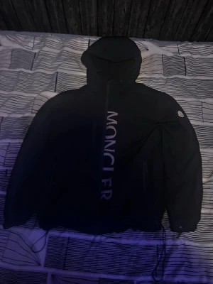 Svart Moncler windbreaker  - Svart vindjacka från Moncler med stor logotyp vertikalt längs dragkedjan och klassisk Moncler-patch på ärmen. Jackan har huva, dragkedjeförsedda fickor och justerbar nederkant. Perfekt för en stilren och sportig look.