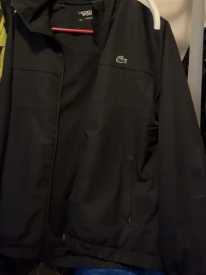 Svart vindjacka från Lacoste Sport - Snygg svart vindjacka från Lacoste Sport med klassisk grön krokodil-logga på bröstet. Jackan har dragkedja framtill, två sidofickor med dragkedja och en hög krage. Perfekt för dig som vill ha en stilren och sportig look.