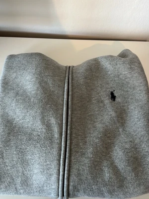 Grå hoodie från Polo Ralph Lauren - Snygg grå zip-hoodie från Polo Ralph Lauren med klassisk broderad logga på bröstet. Hoodien har dragkedja, mjukt innerfoder och är tillverkad i bomullsblandning. Perfekt för en chill och stilren look.