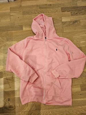 Rosa hoodie från Ralph Lauren - Snygg ljusrosa hoodie från Ralph Lauren med dragkedja och klassisk huva. Broderad logga i mörkblått på bröstet och två fickor framtill. Mjuk bomullskänsla och ribbade muddar vid ärmslut och nederkant. Perfekt för en chill och avslappnad stil. OBS!Felmärkt storlek, men passar för storlek S. Ej använt. Passar för både tjejer och killar