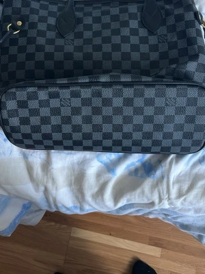 Louis Vuitton Damier Ebene handväska - Snygg handväska från Louis Vuitton i det klassiska Damier Ebene-mönstret. Väskan är svart och mörkbrun med rutigt tryck, har svarta handtag och guldfärgade metalldetaljer. Tillverkad i canvas med läderdetaljer och röd insida. Perfekt för dig som gillar lyxig och tidlös design.
