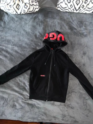 Hugo boss hoodie - Svart hoodie från HUGO med dragkedja och stor röd logga på huvan. HUGO-logga även på bröstet. Klassisk passform med känguruficka och justerbar dragsko i huvan. Mjuk bomullsblandning som känns skön mot huden.