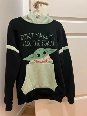 Svart hoodie med Baby Yoda och öron - Svart hoodie med ljusgrön huva och stora Baby Yoda-öron. Framsidan har ett tryck av Baby Yoda och texten 'Don't make me use the force!'. Stor ficka framtill och ljusgröna detaljer på ärmarna. Perfekt för Star Wars-fans som vill sticka ut. Juniorstorlek men är mer som en S. Katt finns i hemmet.