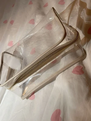 Transparent necessär med vit dragkedja - Säljer en stilren, genomskinlig necessär i plast med vita kanter och dragkedja. Perfekt för att organisera smink eller småprylar. Enkel, rektangulär form och lätt att ta med sig i väskan.