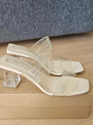 Transparenta sandaler med klack - Säljer ett par trendiga sandaler från Millennial Collection med transparenta remmar och fyrkantig tå. Klacken är också genomskinlig och blockformad, vilket ger en modern vibe. Sulan är ljusbeige och sandalerna har en stilren, minimalistisk look. Klack ca 6 cm 