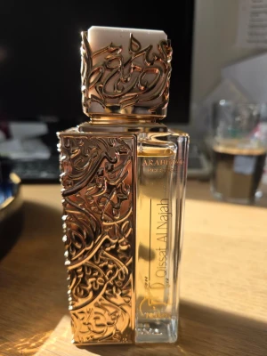 Arabiyat Prestige Dossat Al Najah parfym - Lyxig parfymflaska från Arabiyat Prestige, modell Dossat Al Najah. Flaskan är genomskinlig med detaljer i guld och har ett unikt orientaliskt mönster i relief på både kork och sidor. Perfekt för dig som gillar exklusiva och dekorativa doftflaskor. 