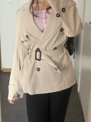 Beige trenchcoat med bälte från vero moda - Stilren beige trenchcoat från vero moda med dubbelknäppning och bälte i midjan. Jackan har stora slag, svarta knappar och justerbara ärmslut. Perfekt för dig som vill ha en klassisk och trendig look. Köpt för 600kr, säljer för 350kr bara! Den är ny, fick den nyligen. Materialet känns mjukt och följsamt. Bältet ingår inte tyvärr.