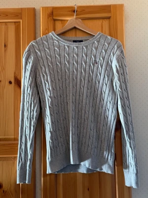 Ljusgrå kabelstickad tröja Gant - Snygg ljusgrå kabelstickad tröja från Gant med rund halsringning och diskret broderad logga på bröstet. Tröjan har långa ärmar och ribbade muddar vid ärmslut och nederkant. Perfekt för lager-på-lager och klassisk stil.