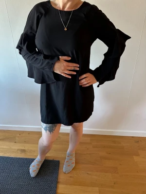 Svart klänning med volangärmar - Svart kort klänning med långa volangärmar och öppen rygg. Klänningen har en enkel, rak passform och är tillverkad i ett mjukt tyg som ger en snygg siluett. Perfekt för dig som gillar stilrena plagg med en twist.