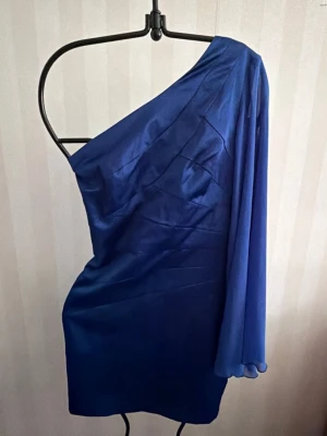 Blå one shoulder kortklänning - Snygg blå kortklänning med one shoulder-design och lång ärm på ena sidan. Klänningen har en figurnära passform och är tillverkad i ett glansigt material som ger en lyxig känsla. Perfekt för dig som vill sticka ut med en unik och trendig look.