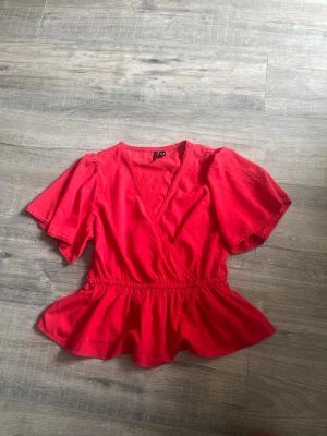 Röd omlottblus med volang - Snygg röd omlottblus med korta puffärmar och volang nertill. Blusen har markerad midja och v-ringning, perfekt nu till sommaren. Köpt från vero Moda 