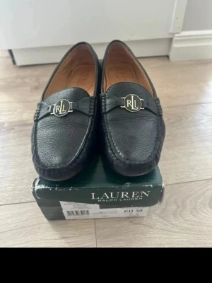 Svarta loafers från Ralph Lauren - Stilrena svarta loafers från Lauren Ralph Lauren i äkta läder med klassisk rund tå och guldfärgad RL-detalj framtill. Skorna har en tidlös design och är perfekta för dig som gillar en clean och elegant look. Äkta läder storlek 39