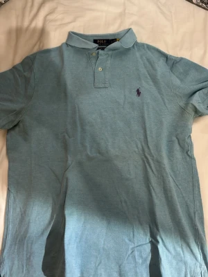 Blå pikétröja från Polo Ralph Lauren - Klassisk ljusblå pikétröja från Polo Ralph Lauren med korta ärmar och krage. Tröjan har två knappar vid halsen och den ikoniska lilla loggan broderad i lila på bröstet. Perfekt för en avslappnad och stilren look.