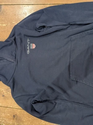 Mörkblå hoodie från GANT - Mörkblå hoodie från GANT med broderad logga och sköld på bröstet. Klassisk känguruficka framtill och huva. Tillverkad i mjukt bomullsmaterial som känns skönt mot huden. Perfekt för en avslappnad och stilren look.