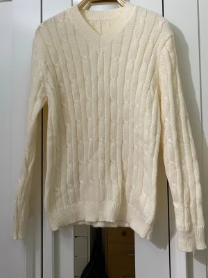 Vit kabelstickad tröja med V-ringning - Kabelstickad tröja i mjuk vit färg med V-ringad hals och ribbade muddar vid ärmslut och nederkant. Klassisk och stilren design som passar perfekt till många olika outfits. Materialet ser ut att vara bomull eller en bomullsblandning.