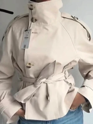 Beige trenchcoat - Stilren beige trenchcoat från bershka med hög krage, knappar framtill och knytskärp i midjan. Jackan har axelklaffar och dekorativa detaljer vid axlarna. Perfekt för en trendig och klassisk look.