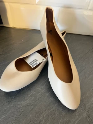 Beige  ballerinaskor från H&M - Stilrena beige ballerinaskor från H&M med rund tå och låg klack. Skorna har en minimalistisk design och är tillverkade i ett slätt syntetmaterial med beige innersula. Perfekta för en clean och enkel look.