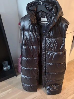 Svart lång dunväst från H&M - Snygg svart lång dunväst från H&M med glansig finish och stor huva. Västen har breda kanaler, dragkedja framtill och praktiska fickor på sidorna. Perfekt för lager på lager och en cool streetstyle-look.