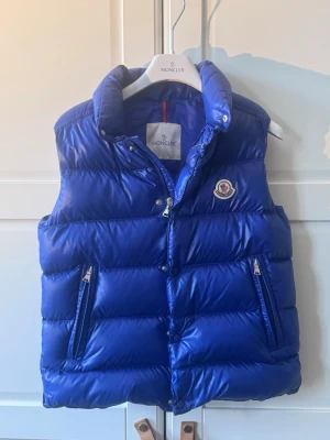 Blå dunväst från Moncler - Snygg blå dunväst från Moncler med glansig finish, hög krage och dragkedja framtill. Västen har två fickor med dragkedja och klassisk Moncler-logga på bröstet. Insidan har ett serietidningsmönster och märkestaggar. Perfekt för lager-på-lager-stil.