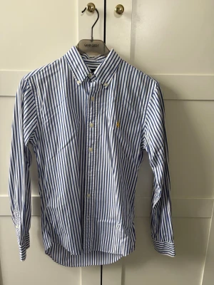 Blåvit randig skjorta från Ralph Lauren - Snygg blåvit randig skjorta från Ralph Lauren i slim fit-modell. Skjortan har klassisk krage, knappar framtill och broderad logga på bröstet. Tillverkad i bomull och har långa ärmar. Perfekt för en stilren look.