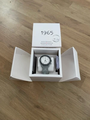 Omega x Swatch Mission To The Moon 1965 - Snygg Omega x Swatch MoonSwatch från Bioceramic MoonSwatch Collection. Klockan har en grå boett och armband med vit urtavla och svarta detaljer. Tydlig tachymeter på ringen och Omega-logga på armbandet. Perfekt för dig som gillar stilrena accessoarer.