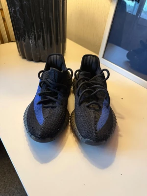 Yeezy Boost 350  - Säljer ett par Adidas Yeezy Boost 350 V2 i svart med blå stripe och texten 'SPLY-350' på sidan. Skorna har en stickad ovandel i mesh och en tjock, räfflad sula i gummi. Perfekta för dig som gillar streetwear och vill sticka ut.
