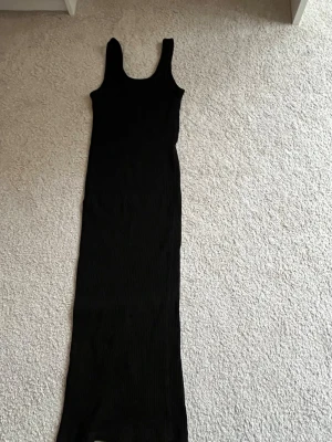 Svart ribbad långklänning H&M - Svart ribbad långklänning från H&M med ärmlös design och rund halsringning. Klänningen har en kroppsnära passform och är tillverkad i ett stretchigt material som ger en snygg siluett. Perfekt för dig som gillar minimalistisk och stilren look.