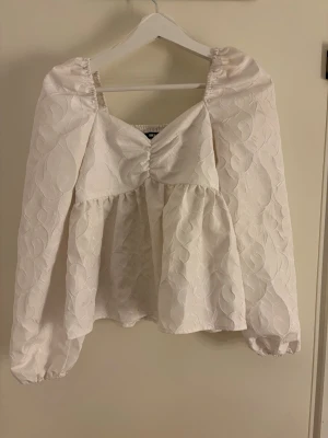 Vit puffärmad blus med struktur - Supersöt vit blus med puffärmar och rynkdetalj framtill. Blusen har ett strukturerat mönster i tyget och smock i ryggen för skön passform. Långa ärmar med resår vid ärmslut och lätt utsvängd nederdel. Perfekt för en trendig look.