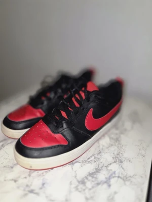 Nike Court Borough Low 2 Sneakers, Svart/Röd, EU 36 - Snygga sneakers från Nike i svart och rött med klassisk swoosh-logga på sidan. Skorna har lågt skaft, svarta skosnören och vit sula. Ovandelen är i läder med perforerade detaljer på tån för extra stilpoäng.