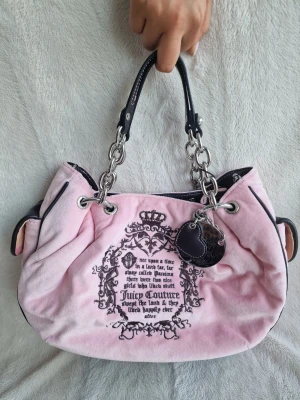 Rosa Juicy Couture handväska i velour - Söt rosa handväska från Juicy Couture i mjuk velour med svarta detaljer och silverfärgade kedjor. Väskan har tryckt text och emblem på framsidan samt ett hjärtformat hänge. Perfekt för dig som gillar en trendig och lekfull stil.