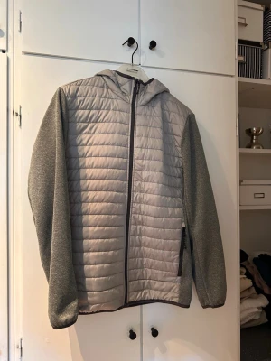 Grå vindjacka med huva - Snygg grå vindjacka med quiltad framsida och rygg samt mjuka, melerade ärmar. Jackan har hel dragkedja, två sidofickor med dragkedja och en praktisk huva. Perfekt för blåsiga dagar och enkel att matcha med olika outfits. Pytte litet hål på magen! 