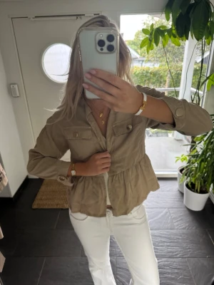 Beige overshirt med volangdetalj - Snygg beige overshirt med volangdetalj nedtill och två bröstfickor med knappar. Jackan har lång ärm och stängs med knappar framtill. Perfekt att slänga över en topp för en avslappnad men trendig look.