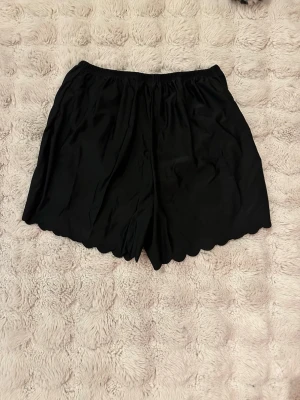 Oanvända Svarta  simshorts med vågig kant - Svarta shorts med elastisk midja och snygg vågig kant nertill. Tillverkade i ett mjukt och lätt material som ger en skön känsla. Perfekta för varma dagar och enkel att matcha med olika toppar.