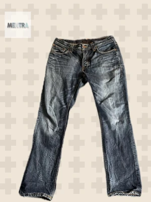 Blå jeans från Nudie Jeans - Säljer ett par klassiska blå jeans från Nudie Jeans med snygga slitningar och kontrastsömmar. Jeansen har fem fickor, raka ben och detaljerade bakfickor med Nudie-signatur. Tillverkade i slitstark denim, perfekta för en avslappnad stil.