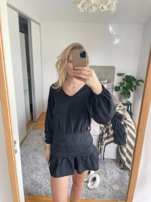 Svart kort klänning med v-ringning - Svart klänning med v-ringning och långa ärmar. Modellen är kort med en markerad midja och volang nedtill. Klänningen har en loose passning och är tillverkad i ett lätt tyg, perfekt för en avslappnad och trendig look. Den är omsydd i kragen 