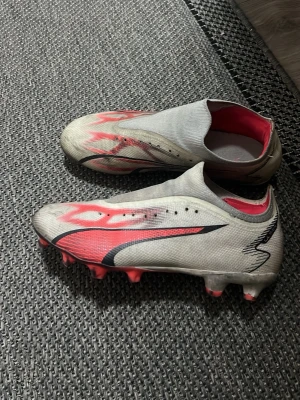 Puma Ultra fotbollsskor vit/röd - Säljer ett par snabba Puma Ultra fotbollsskor i vitt med röda och svarta detaljer. Skorna har låg vikt, strumpliknande passform och mönstrad ovandel i syntetmaterial. Perfekta för dig som vill ha explosiv fart på planen och snygg design.