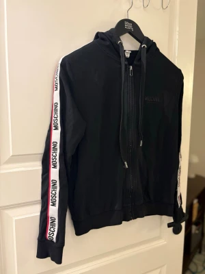 Svart hoodie från Moschino med logoband - Svart hoodie från Moschino med dragkedja framtill och huva med snören. Ärmarna har vita logoband med svart Moschino-tryck och en röd linje längs sidorna. Ribbstickade muddar vid ärmslut och nederkant. Perfekt för en trendig streetstyle-look. En söm har gått ut syns i en av bilderna men inget som syns när man har på sig koftan xs-S 
