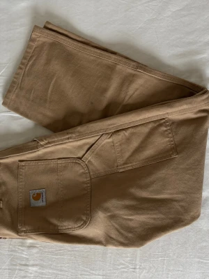 Bruna double knees från Carhartt - Säljer ett par klassiska bruna cargobyxor från Carhartt med stora fickor på sidorna och robust bomullsmaterial. Perfekta för dig som gillar streetwear och funktionella detaljer. Byxorna har en avslappnad passform och Carhartt-logga på fickan.